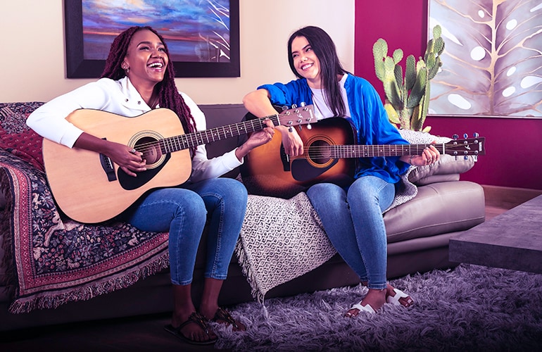 Two women smiling on a couch while playing F310 acoustic guitars in Natural and Tobacco Brown Sunburst.